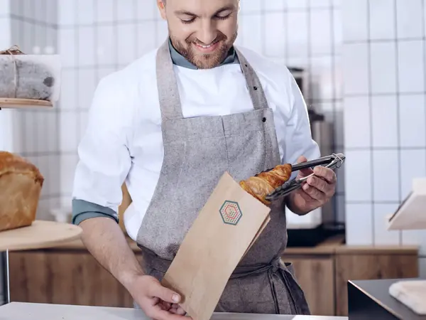 In einer Bäckerei verpackt ein Bäcker Backwaren in einem Faltenbeutel
