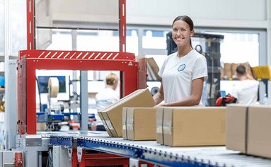 Eine Mitarbeiterin aus der Logistik steht an einer Packstraße und hält ein gepacktes Paket in der Hand.