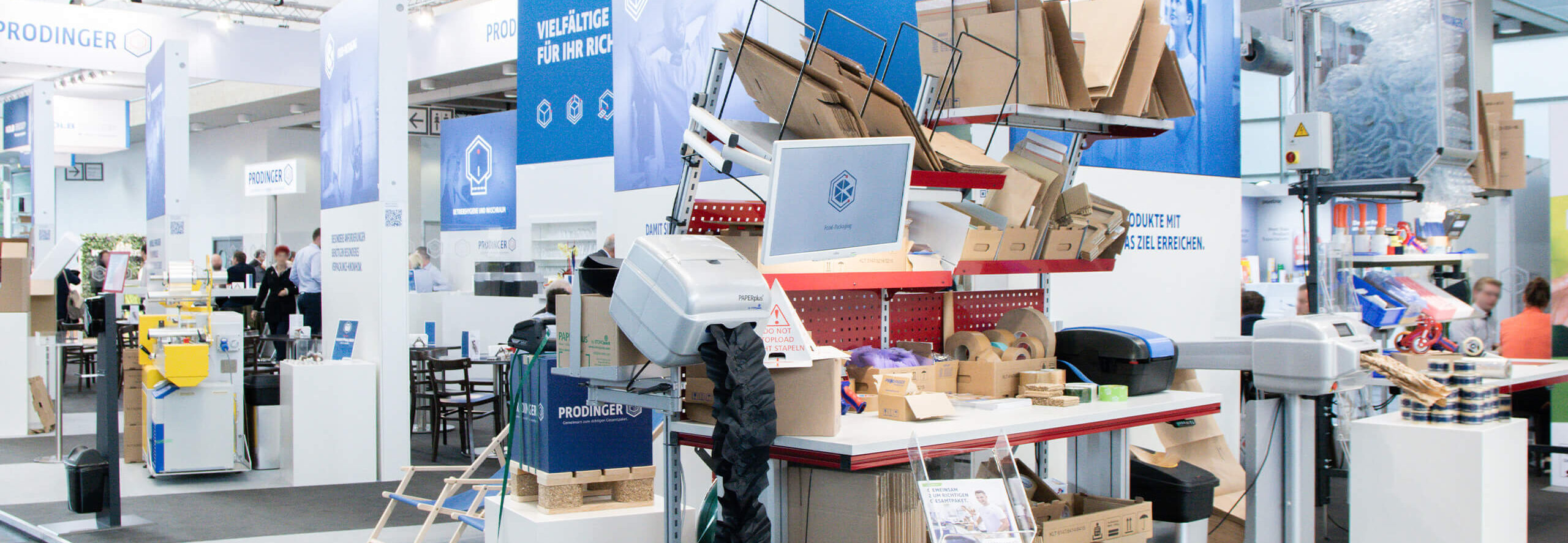 Messestand von PRODINGER Verpackung, auf der ein Packtisch mit Polstermaschine, Klebebänder und verschiedene Kartons steht. 