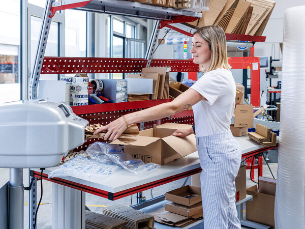 Mitarbeiterin steht an einem Packtisch und füllt einen Karton mit Polstermaterial.